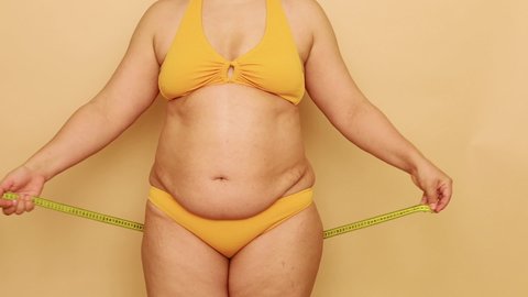 Unrecognizable overweight woman standing in yellow bra, bikini, showing excess belly, measuring buttocks with tape on beige background. Body positive, cellulite, obesity, weight control, liposuction.