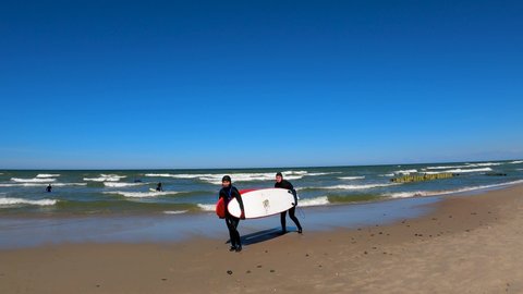 Kaliningrad, Russia, 12, March, 2022:
Two girls in diving suits smile and drag surfboards, two girls with surfboards under their arm after riding in the cold sea