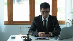 An Asian male lawyer is using a smartphone to consult clients in the office with wooden hammers on courtroom tables and goddess scales with laptop concepts of justice and law. good service consulting