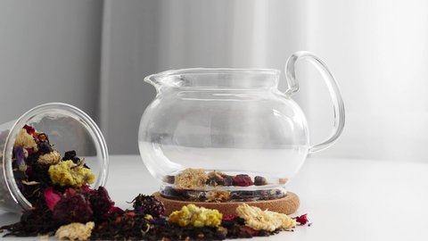 Pour hot water from an electric kettle into a glass teapot with fruit tea. Green tea with fruits is brewed in boiling water. The tea leaves swirl from the movement of water. A spill of tea on the tabl