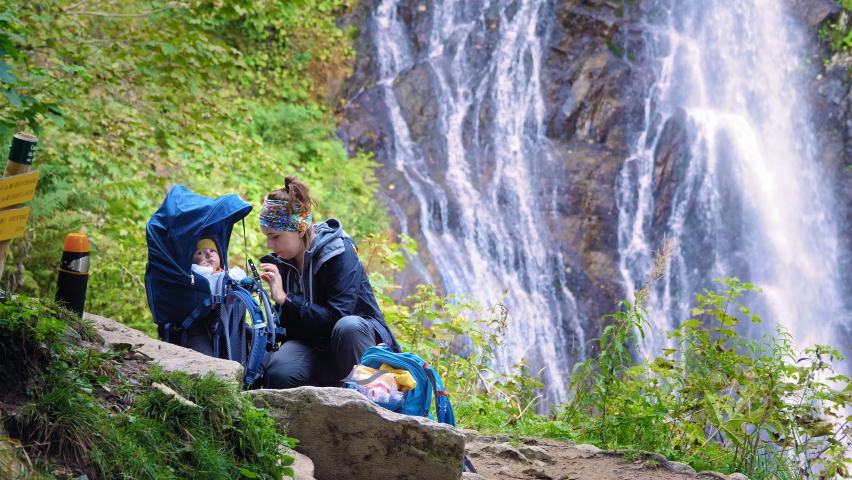 Mother with a baby (in a carrier) on a break during a hike in the beautiful nature. Slow motion.