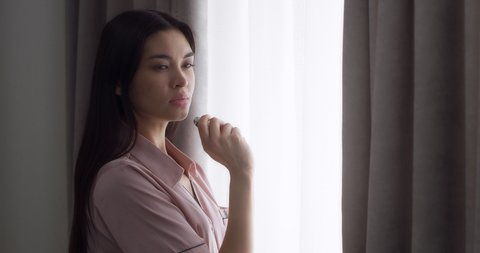 Young woman with long dark hair dressed in pink silk pajamas is standing near the window in a room at home and vaping, smoking an eron cigarette, blowing smoke.
