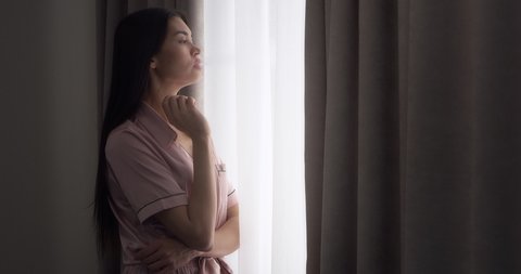 Upwards. Young woman with long dark hair dressed in pink silk pajamas is standing near the window in a room at home and vaping, smoking an eron cigarette, blowing smoke.