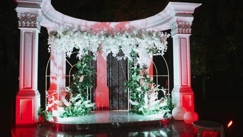 Luxurious wedding arch is decorated with floral arrangements and sequins on threads and illuminated by colorful lights. Night scene. Exquisite wedding arch with columns and frame. Feast day. Romance