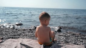 A boy-child is sunbathing, sitting with his back to the camera looking at the sea. Vacation on the seashore with the whole family. Slow motion video