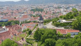Inscription on video. Graz, Austria. Clock Tower in Graz. The historic city center aerial view. Mount Schlossberg (Castle Hill). Arises from blue water, Aerial View, Departure of the camera
