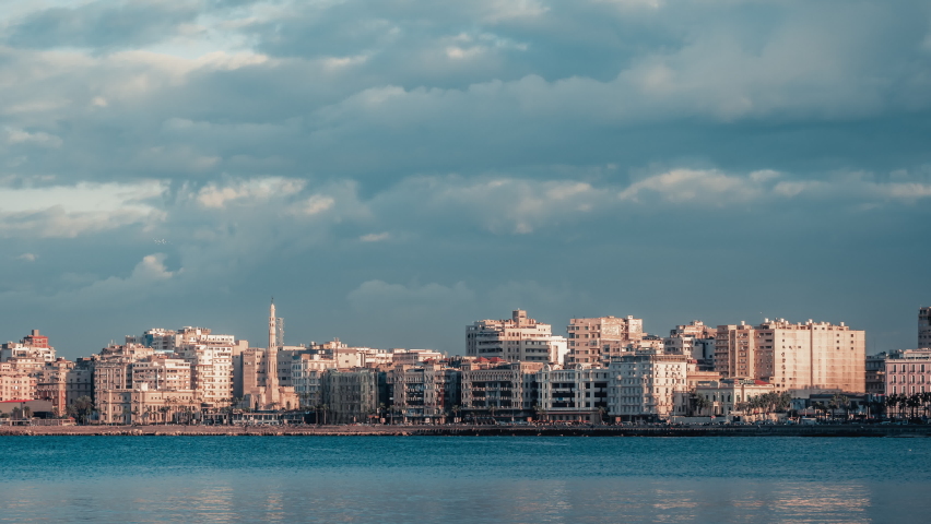 Sea coast of Alexandria. Seashore of ancient city of Egypt with buildings, constructions, palms. Timelapse. Urban view of seaside with houses, trees. Cloudy day