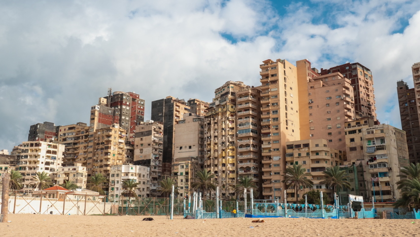 Sea coast of Alexandria. Seashore of ancient city of Egypt with buildings, constructions, palms. Timelapse. Urban view of seaside with houses, moving people, trees. Cloudy day