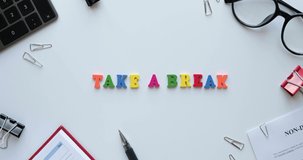Flatlay with office items and phrase Take a break from colored wooden letters. Layout on a white background. Glasses, keyboard, pen, paper clips, diary, work papers. 4K looped stop motion animation