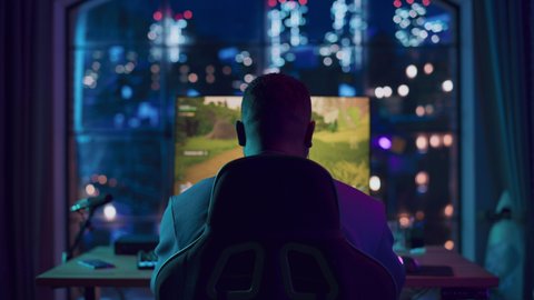 Back of the Head Angle of a Successful African American Gamer Putting on Headphones and Playing a Video Game on Personal Computer in a Neon Lit Living Room at Home. Young Man in Victorious.