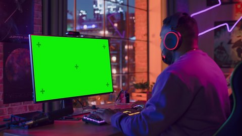 Excited Gamer Playing Online Video Game with a Mock Up Green Screen on His Powerful Personal Computer. Room and PC have Colorful Neon Led Lights. Cozy Evening at Home in Loft Apartment. Static Shot.
