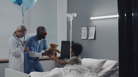 Locked-down of boy with oxygen tube in nose lying on bed, then sitting up when female doctor and male nurse with clown nose walking in and giving air balloons and toy to kid