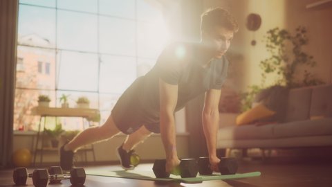Fit Athletic Strong Young Man Performing Enduring Training in Plank Position, while Lifting Dumbbells During Morning Workout at Home in Sunny Apartment. Concept of Healthy Lifestyle and Fitness.