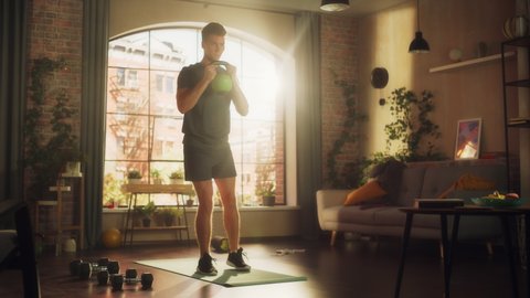 Strong Athletic Fit Young Man Lifting and Squatting with a Heavy Kettlebell, Doing Core Strengthening Exercises During Morning Workout at Home in Sunny Apartment. Concept of Health and Fitness.