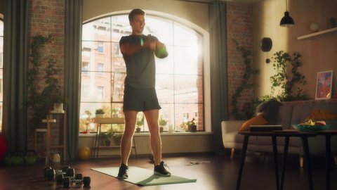 Motivated Athletic Fit Young Man Swinging a Heavy Kettlebell from Hand to Hand, Doing Core Strengthening Exercises During Morning Workout at Home in Sunny Apartment. Concept of Health and Fitness.