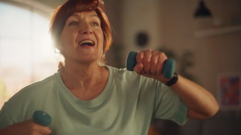 Portrait of a Middle Aged Female Exercising and Training at Home on Sunny Morning. Elderly Female Strengthening Arm Muscles with Dumbbell Workout in Loft Apartment Living Room. Wellness and Fitness.