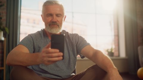 Close Up Portrait of a Fit Middle Aged Man Using Smartphone in Between Workouts During Morning Exercises at Home in Sunny Apartment. Healthy Lifestyle, Fitness, Recreation, Wellbeing and Retirement.