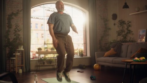 Strong Athletic Fit Senior Man Doing Cardio High Knees and Core Strengthening Exercises During Morning Workout at Home in Sunny Apartment. Fitness and Recreation Concept. Zoom Out Shot.