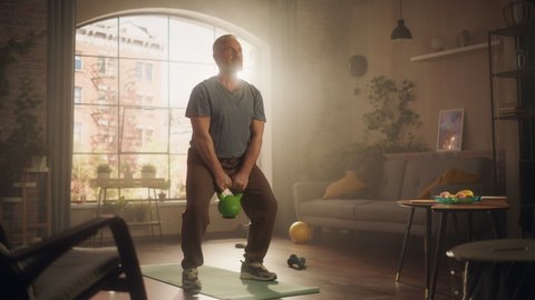 Strong Athletic Fit Senior Man Lifting and Swinging a Heavy Kettlebell, Doing Core Strengthening Exercises During Morning Workout at Home in Sunny Apartment. Concept of Retirement and Fitness.