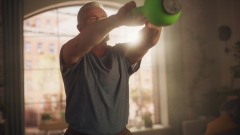 Portrait of a Strong Fit Senior Man Lifting and Swinging a Heavy Kettlebell, Doing Core Strengthening Exercises During Morning Workout at Home in Sunny Apartment. Concept of Wellness and Fitness.