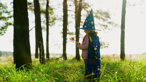 Girl, child plays in wizard costume, holds toy with a magic wand in park. Family walk. Halloween. Happy childhood, family. Children's fantasies in wizard's robes in forest. Kid plays magician outdoors