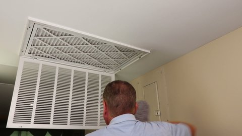 Guy changing a dirty furnace air filter with a clean one and dusting inside and outside the vent intake grate. Male replacing an old air filter with a new one in a home furnace and dusting vent door