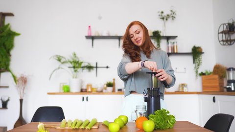 A woman with red hair stands in her kitchen and makes celery juice with a juicer
