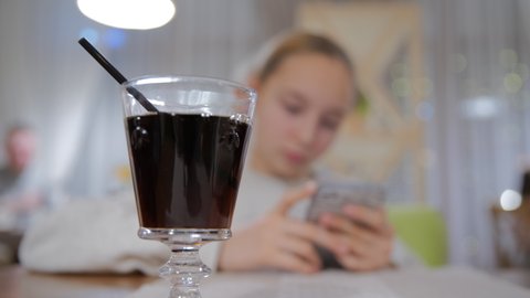 Close-up glass of coke with black straw selective focus. Soft drink on table in cafe. Blurred girl with mobile phone on background. DOF