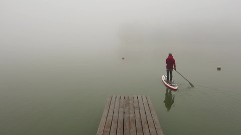 Khmelnytskyi - 12.03.2020: man stand up paddleboard board floats water river in thick fog. man sails away from wooden pier. extreme water sport stand-up paddle surfing 4k aerial top view drone ukraine