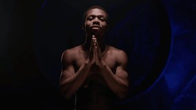 Cool African American teenager with bare, pumped-up body sings hip-hop song and emotionally gestures with his hands. Man raps in music video and sings rap song against blue underground background. 