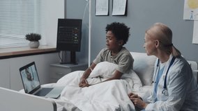 Medium of Black boy with oxygen tube in nose sitting up on bed with blond female Caucasian doctor in hospital room at daytime, video calling little girl and her dad via laptop computer