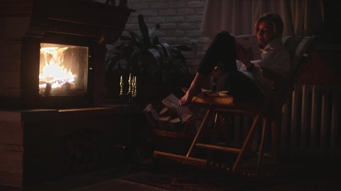 A girl is sitting in a rocking chair and reading a book near the fireplace. Smiling in a cozy room. High quality 4k footage