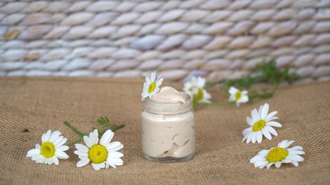 Close up homemade natural eye cream with chamomile flowers in transparent glass jar on beige background.