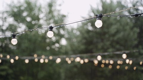 Heavy rain falls on vintage garlands strung on street near trees with turned on round bulbs. Drops of water hang from electric wires and glass bulbs of light bulbs.