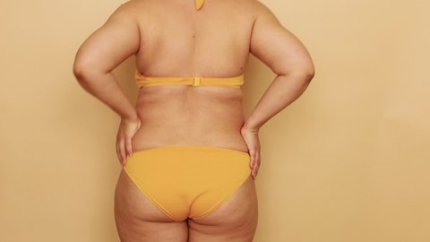 Back view of unrecognizable fat woman wearing yellow bra, bikini, dancing, making active moves, putting hands waist, moving buttocks on beige background. Body positive, dance, weight loss, having fun.