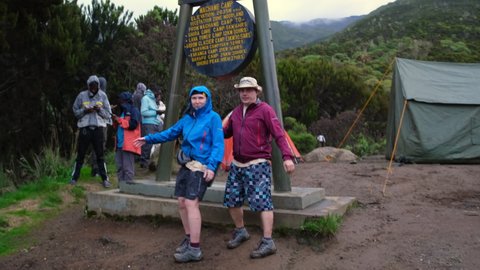 Kilimanjaro. Tanzania. Africa - 12.24.2021 Two hikers are photographed against the stele at Machame camp 2835 m while climbing Mount Killimonjaro with mountain views and tents