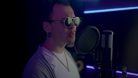 Professional singer records new songs in studio with blue led light. Bearded man in sunglasses heartily sings into black microphone closeup