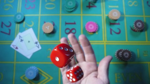 Male hand tossing up red dice in slow motion against background of green roulette table in casino. Man gambler, gambling, craps, poker. Casino chips and cards are laid out on gaming table close up.