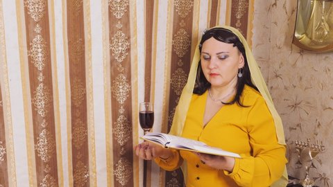 A Jewish woman in a wig and head covering makes a celebratory kdush with a glass in her hand and a siddur.
