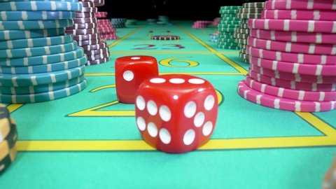 Camera pans over green gaming table with stacks of casino chips on a black background. Red dice thrown on the table fly straight into camera. Craps. Concept of betting. Close up in slow motion.