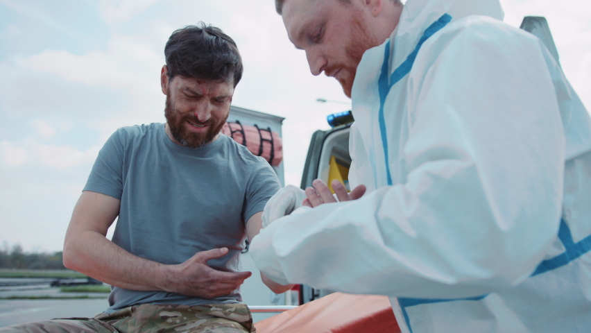 Outdoors medic provides first aid and rewinds his hand with a bandage to wounded soldiers scream in pain near ambulance. Military concept. War