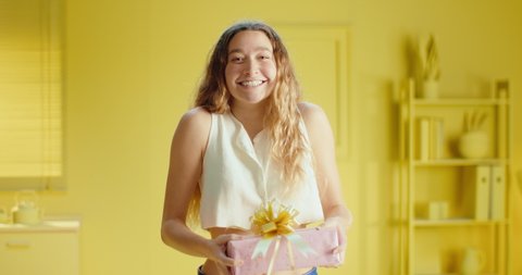 Slow motion of a close up joyful Caucasian woman in sleeveless clothes holding a gift box and looking at camera while smiling and small jump in the studio with monochrome yellow color interior.