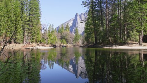 Scenic peaks reflected in clear water surrounded by green pine forest. Cathedral Rocks in Yosemite valley reflecting in Merced River with glacier water slowly flowing with rippling lake like mirror