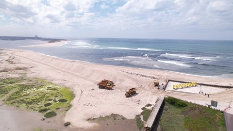 Esposende, Portugal, April 10, 2022: DRONE AERIAL FOOTAGE - The two sides of Restinga de Ofir. Esposende fishing harbor maintenance dredging. Dumper vehicles and rotating excavator parked on the sand.