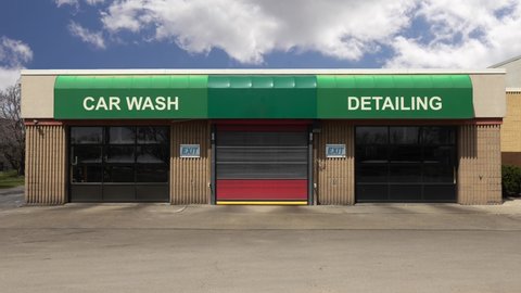 Clouds in a blue sky floating over car wash building. Three garage doors at the exit. Green awning stating car wash. one red garage door. Concrete building. Security camera on building. 