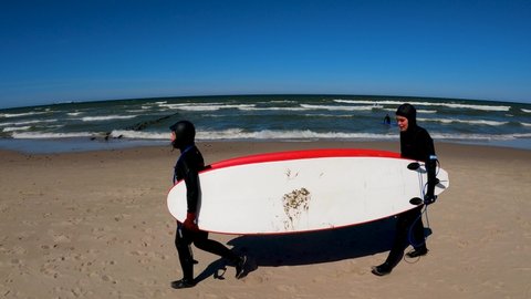 Kaliningrad, Russia, 12, March, 2022:
Two girls in diving suits dragging surfboards, two girls with surfboards under their arm after riding in the cold sea