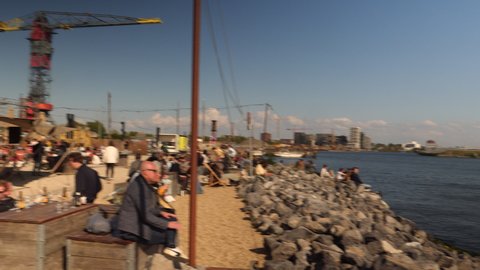 Amsterdam, Netherlands, April 25, 2022. Lounge bar on the waterfront crowded with people on a sunny day. Pan from bar to river. 