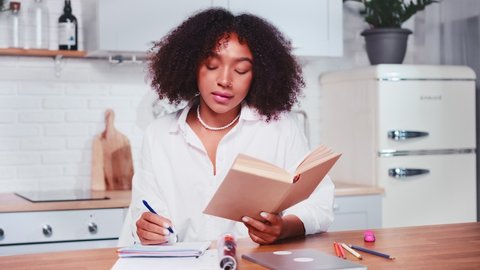 Young attractive African American woman student making notes in workbook holding textbook in hands preparing for test at university sits at table in kitchen. College, education, school
