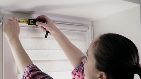 A handy woman with a tape measure installs white roller blinds indoors. Shutters day and night