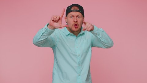 Man showing loser gesture and pointing on you, blaming accusing for unsuccess, expressing disrespect, mocking your failure, bullying abuse. Young guy isolated alone on pink studio wall background